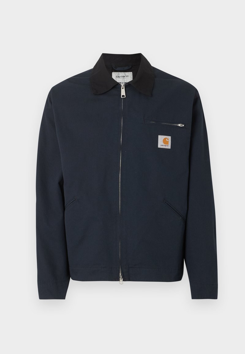 Donkerblauw jack met rits, zwarte kraag, twee schuine zakken aan de voorkant en een kleine met rits afsluitbare borstzak met een Carhartt-logo patch.