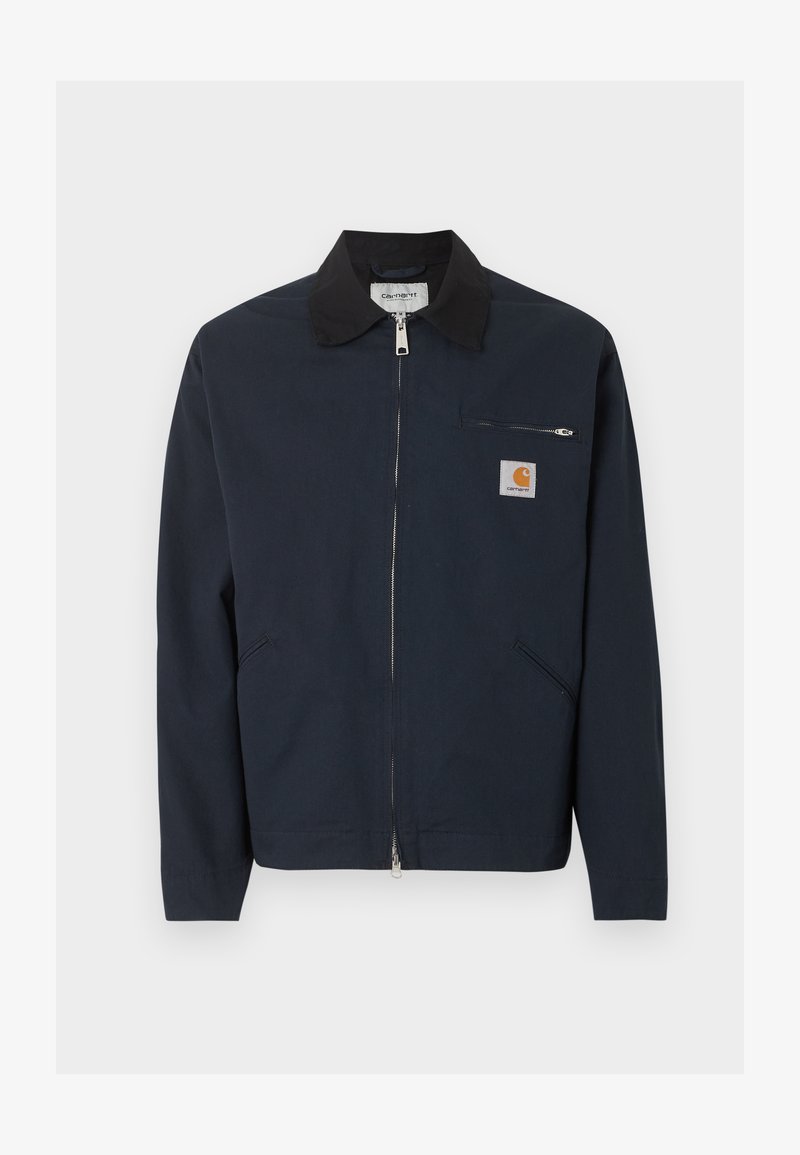 Dunkelblaue Zip-up-Jacke mit schwarzem Kragen, zwei schrägen Fronttaschen und einer kleinen Reißverschlusstasche auf der Brust mit einem Carhartt-Logo-Patch.