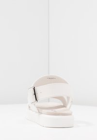 Bílé kožené sandály s texturovaným povrchem, nastavitelným páskem a silnou bílou podrážkou. Design obsahuje minimalistické linie a akcenty.