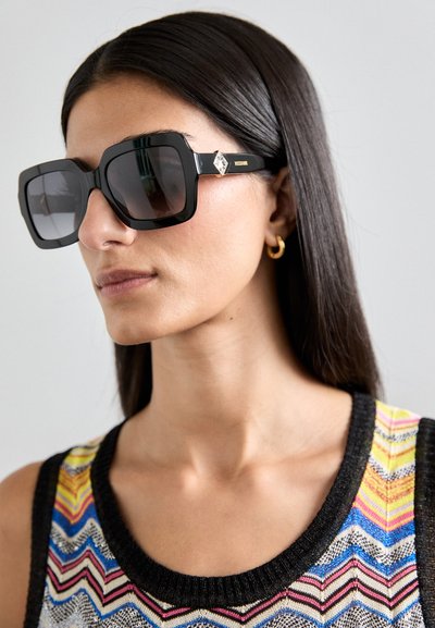 Gafas de sol negras de gran tamaño con un marco geométrico, lentes oscuros y un elemento decorativo en la varilla, combinadas con una blusa de patrones multicolores.
