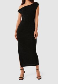 Svart enaxlad klänning med en figurnära design, strukturerat tyg och en sidoflik. Buren med svarta sandaler med remmar.