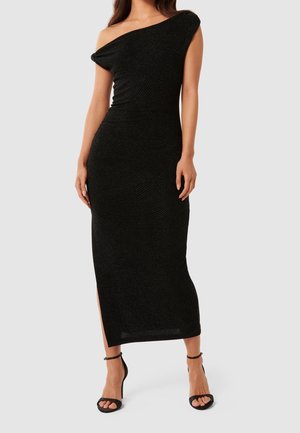 Robe noire à une épaule avec un design ajusté, en tissu texturé et une fente sur le côté. Portée avec des talons noirs à lanières.