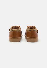 Zapatillas de cuero marrón con un cuello de textil estampado, punta redonda y suela de goma plana con agarre texturizado. La vista trasera muestra detalles de los cordones.