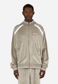 Taupe színű kordbársony zipzáras dzseki fehér részletekkel, rugalmas mandzsettákkal és magas gallérral. A mellkason logó található, valamint oldalsó zsebek.