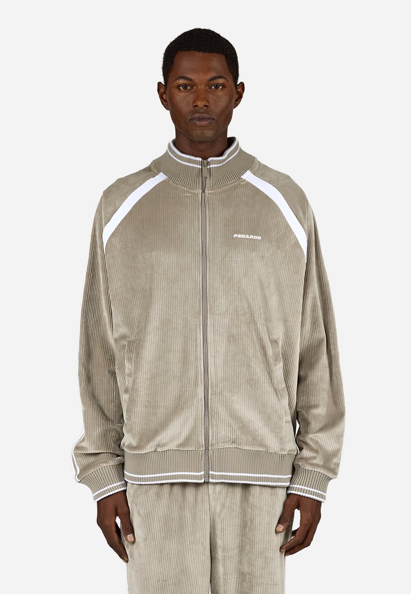 Taupe színű kordbársony zipzáras dzseki fehér részletekkel, rugalmas mandzsettákkal és magas gallérral. A mellkason logó található, valamint oldalsó zsebek.