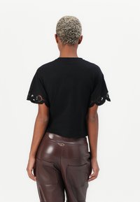Černý crop top s propracovaným krajkovým volánem na rukávech, vyrobený z měkké tkaniny. V kombinaci s lesklými hnědými kalhotami z umělé kůže.
