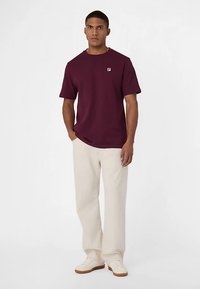 Maglietta bordeaux a maniche corte con un piccolo logo sul petto, abbinata a pantaloni beige a gamba larga e sneakers bianche. Design semplice e casual.