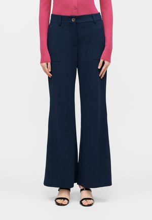 Γυναίκα που φοράει μπλε σκούρο παντελόνι φαρδιάς γραμμής με μπροστινές τσέπες, συνδυασμένο με ροζ μακρυμάνικη μπλούζα και μαύρα σανδάλια ανοιχτούς δακτύλους.