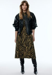 Schwarze Lederbomberjacke über einem langen, braunen Kleid mit einem schwarzen Zebra-Streifenmuster. Knielange schwarze Stiefel runden den Look ab.