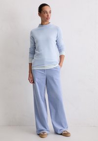 Hellblauer Pullover mit hohem Kragen und gestreiften Bündchen, kombiniert mit weit geschnittenen, gestreiften blauen Hosen und beigefarbenen Schuhen.