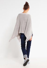 Poncho grigio chiaro con frange e righe orizzontali, indossato sopra una maglietta grigia e jeans scuri. Il materiale sembra tessuto con una finitura testurizzata.