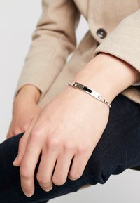 Pulsera de plata con un charm rectangular liso y eslabones de cadena entrelazados, llevada en una muñeca con una chaqueta clara de fondo.