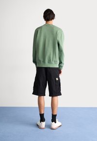 Green sweatshirt with a crew neck, paired with black cargo shorts. The shorts feature side pockets and a logo patch. White athletic shoes.