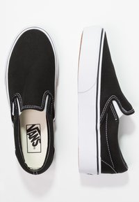 Zapatillas de lona negras sin cordones con suelas de goma blanca y costuras. Presentan acentos elásticos en los lados y un cuello acolchado para mayor comodidad.