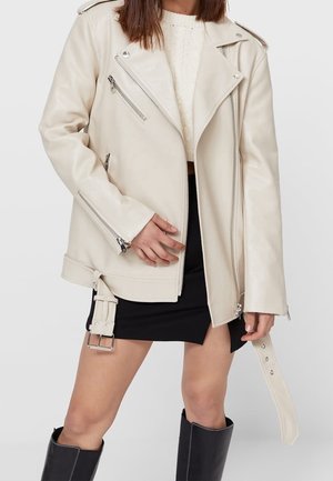 Woman wearing a cream leather jacket with zipper details, a white knit top, a black skirt, and black knee-high boots, standing against a white background.