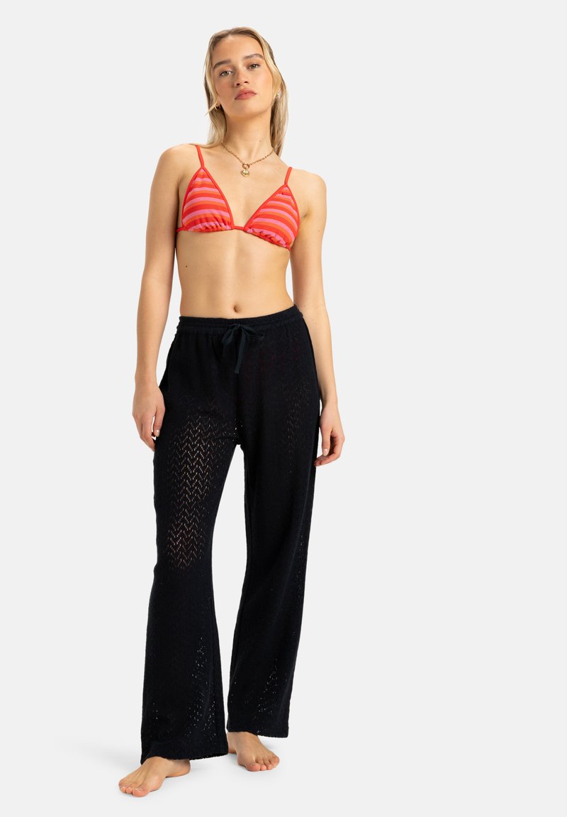 Pantalones de crochet negros con un patrón y cintura con cordón, combinados con una parte superior de bikini a rayas rojas y rosas. La modelo está descalza contra un fondo blanco.