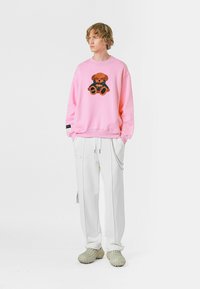 Sudadera rosa claro con un gráfico de oso texturizado, pantalones blancos y zapatos verdes gruesos, con detalles como rayas y cremalleras.