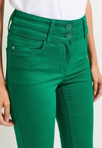 Ripresa ravvicinata di una persona che indossa jeans verdi a vita alta con doppio bottone, tasche frontali e passanti per cintura, abbinati a una maglietta bianca.