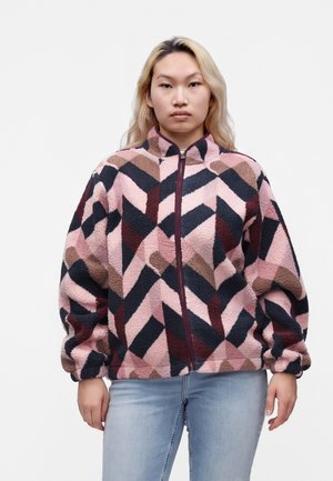 Person med langt blond hår iført en fleecejakke med lynlås med pink, sort og brun geometrisk mønster og lyseblå jeans.