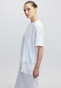 Top blanco de manga corta con un corte holgado, que presenta un diseño de dobladillo en capas y una textura suave. Lleva puestos pantalones blancos a juego.