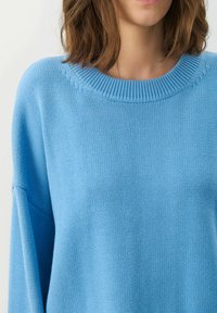 Hellblauer gestrickter Pullover mit einem gerippten Rundhalsausschnitt und überschnittenen Schultern. Die Textur ist weich mit einer glatten Oberfläche.