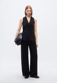 Μαύρο αμάνικο τοπ με βαθύ V ντεκολτέ, σε συνδυασμό με μαύρο παντελόνι φαρδιάς γραμμής. Το μοντέλο κρατά μια μεγάλη, μαλακή μαύρη τσάντα. Ομαλή υφή υφάσματος.