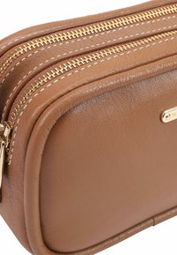 Bruine leren toilettas met goudkleurige ritsen, witte stiksels en een klein logo plaatje. Heeft een ronde vorm en een strak design.