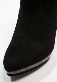 Chaussure en daim noir à bout pointu avec une bande métallique brillante près de la semelle, photographiée sur un fond blanc.