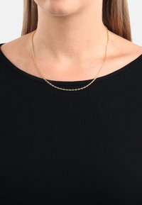 Guldhalsband med sammankopplad oval mönster, vilande p å bar hud, buren över en svart ribbad topp med vid nedskärning.