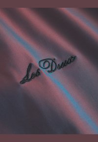 Siden tyg i djup burgundy med iriserande blå ränder; har broderad svart text "Los Deux" i kursiv stil.