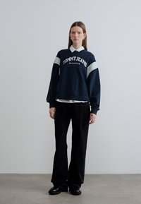 Μπλε σκούρο φούτερ με λευκές ρίγες και το λογότυπο "TOMMY JEANS", φορεμένο πάνω από λευκό πουκάμισο με γιακά, συνδυασμένο με μαύρο παντελόνι φαρδύ.