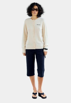 Femme portant un cardigan crème boutonné avec le texte "RÉSUMÉ", pantalon noir court, sandales à plateforme noires, lunettes de soleil et bracelets, debout sur fond blanc.