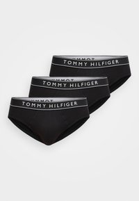 Τρία μαύρα ανδρικά εσώρουχα Tommy Hilfiger με λευκές ελαστικές ζώνες μέσης με το λογότυπο, τοποθετημένα επικαλυπτόμενα σε λευκό φόντο.