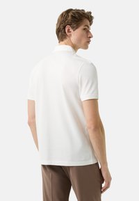 Bílá polo košile s klasickým límcem a krátkými rukávy, hladká textura a boční štěrbiny na lemu, nošená s hnědými kalhotami.