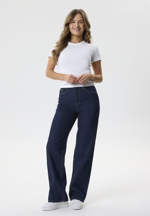 Joven mujer con el pelo largo y ondulado está de pie sonriendo, lleva una camiseta blanca lisa, vaqueros anchos azul oscuro y zapatillas blancas.
