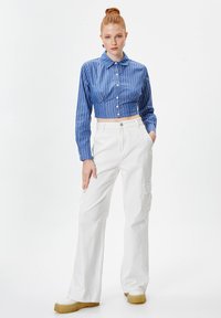 Camicia a righe blu e bianche, corta, con bottoni, abbinata a pantaloni cargo bianchi a vita alta e scarpe con plateau color crema. Tessuto liscio, vestibilità rilassata.