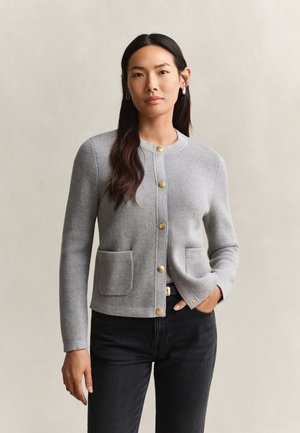 Femme aux longs cheveux foncés portant un cardigan gris clair boutonné avec des poches à l'avant et un pantalon noir, debout devant un fond uni.