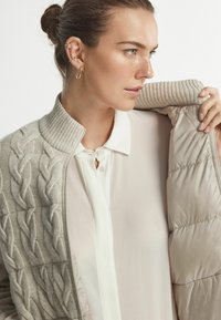 Ljust beige stickad cardigan med ett kabelmönster, ribbad krage och mjukt foder. Under finns en blek skjorta med knappar och nyckelhålsdetalj.