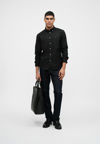 Czarna koszula zapinana na guziki z białymi guzikami, ciemne dżinsy, czarne skórzane buty. Trzyma dużą czarną torbę na ramię. Prosty, elegancki design.