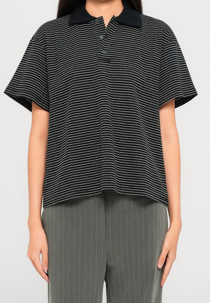 Femme portant un polo à manches courtes rayé noir et blanc avec un col noir, associé à un pantalon gris à fines rayures.