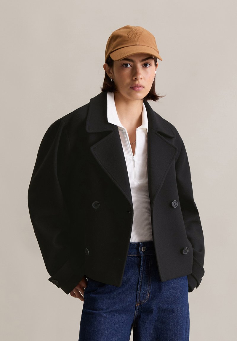 Manteau noir à double boutonnage avec larges revers, en tissu laineux, porté sur un top côtelé blanc et un jean bleu, associé à une casquette de baseball marron.