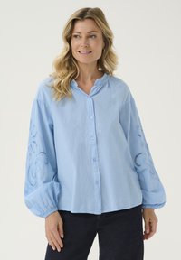 Camicia azzurra chiaro con maniche lunghe a sbuffo e dettagli ricamati. Tessuto morbido con una vestibilità rilassata, colletto arrotondato e yoke arricciato.