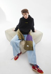 Zwarte hoodie met gele tekst, losse lichtblauwe jeans, olijfgroene en paarse sjaal, rode sneakers met witte accenten, zittend op een textuurster crème ottoman.