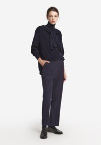 Mörkblå ulltröja med hög krage och draperad scarfdetalj. Skärpda mörka byxor och svarta läderloafers fulländar outfiten.