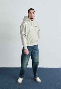 Felpa leggera beige con tasca frontale e cordini, abbinata a jeans blu sbiaditi e sneakers crema. Design semplice con dettaglio del logo.