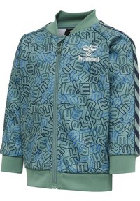 Grön zip-up jacka med ett geometriskt mönster i blått och svart, ribbade ärmar och krage; har en logotyp på vänster bröst och sidofickor.