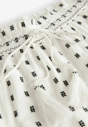 Valkoinen kangas mustilla rusetti- painatuksilla, joustava vyötärö, palmikkonauha ja valkoiset tupsut. Karkea, kevyt materiaali.