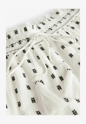 Valkoinen kangas mustilla rusetti- painatuksilla, joustava vyötärö, palmikkonauha ja valkoiset tupsut. Karkea, kevyt materiaali.