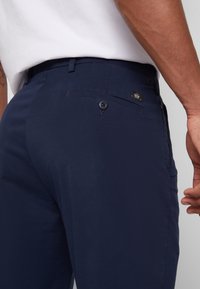 Gros plan d'une personne portant un pantalon bleu marine avec une poche arrière boutonnée et une chemise blanche rentrée.