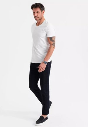 Camiseta blanca de cuello en V, jeans de mezclilla oscuros, zapatos negros tipo slip-on. Presenta un corte masculino y un tatuaje en el brazo superior, posando frente a un fondo neutro.